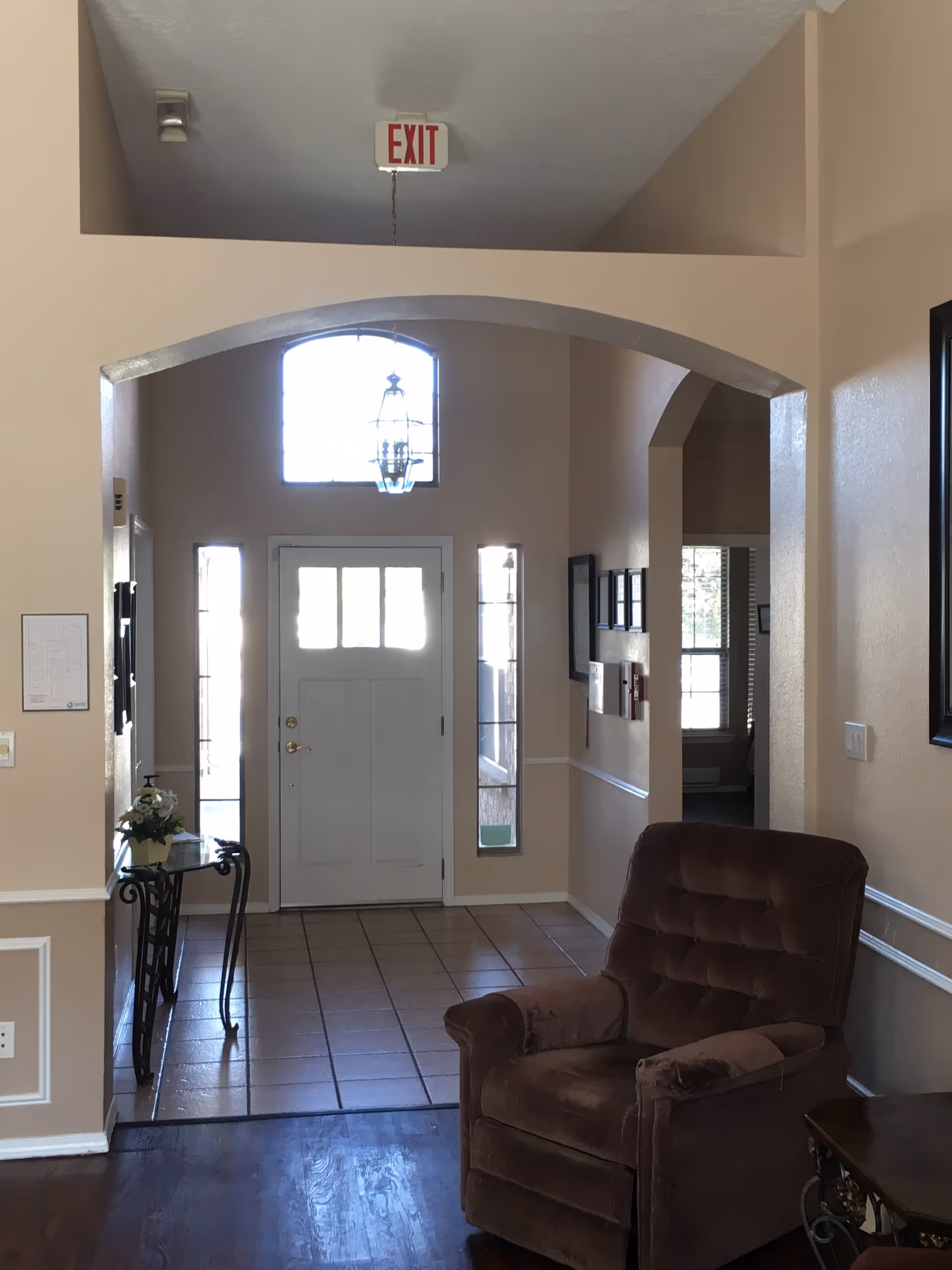 Interior view of an assisted living facility entrance area with a white front door flanked by narrow windows. There is a small glass table with a flower arrangement on the left side and a brown upholstered armchair on the right. The floor near the door is tiled, transitioning to dark wood flooring in the foreground. An exit sign hangs from the ceiling above an archway.