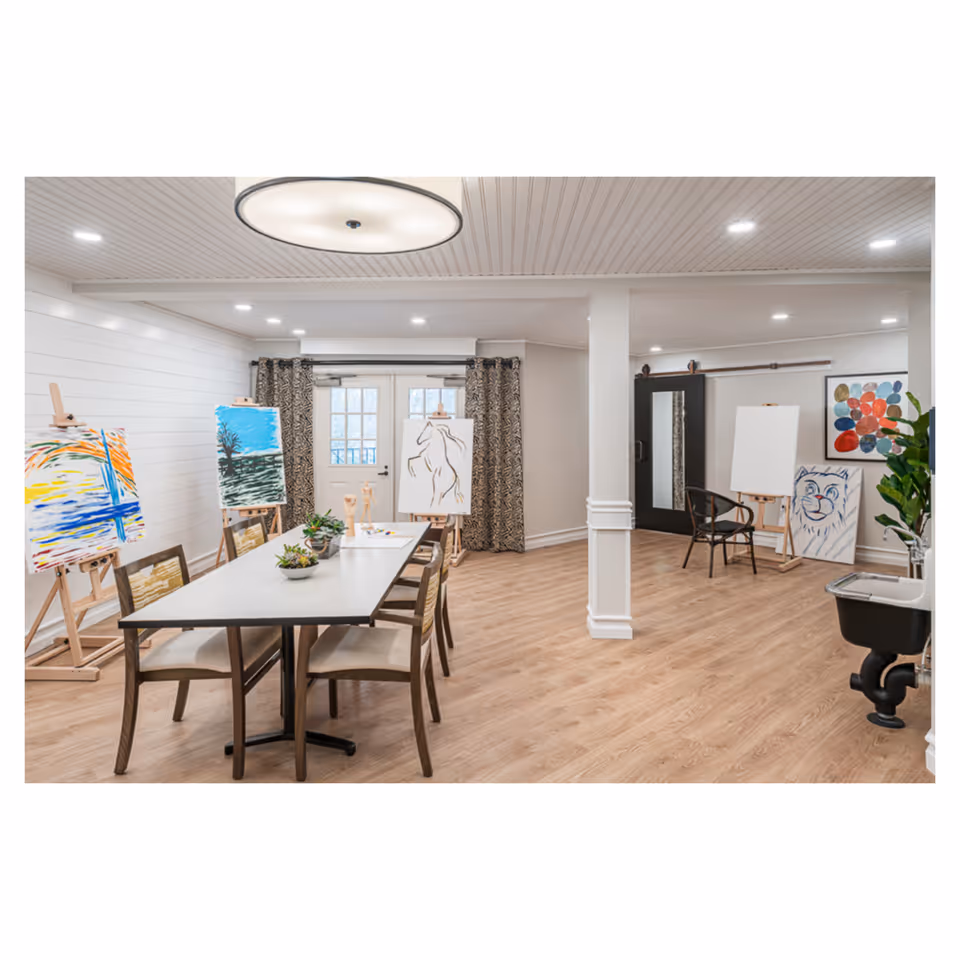 A spacious art room with wooden flooring and white paneled walls. The room features a long table with six chairs around it, small potted plants on the table, and several easels displaying colorful paintings and blank canvases. There is a large circular ceiling light, patterned curtains covering glass doors, a black chair, a black cabinet with a mirror, and a small black sink with a faucet on the right side.