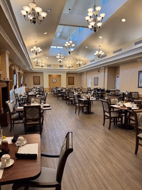 A spacious dining room with multiple tables and chairs arranged neatly. The room features elegant chandeliers hanging from a high ceiling with skylights, beige walls with framed artwork, and a wooden floor. Tables are set with cups, plates, and condiments, creating a welcoming atmosphere.
