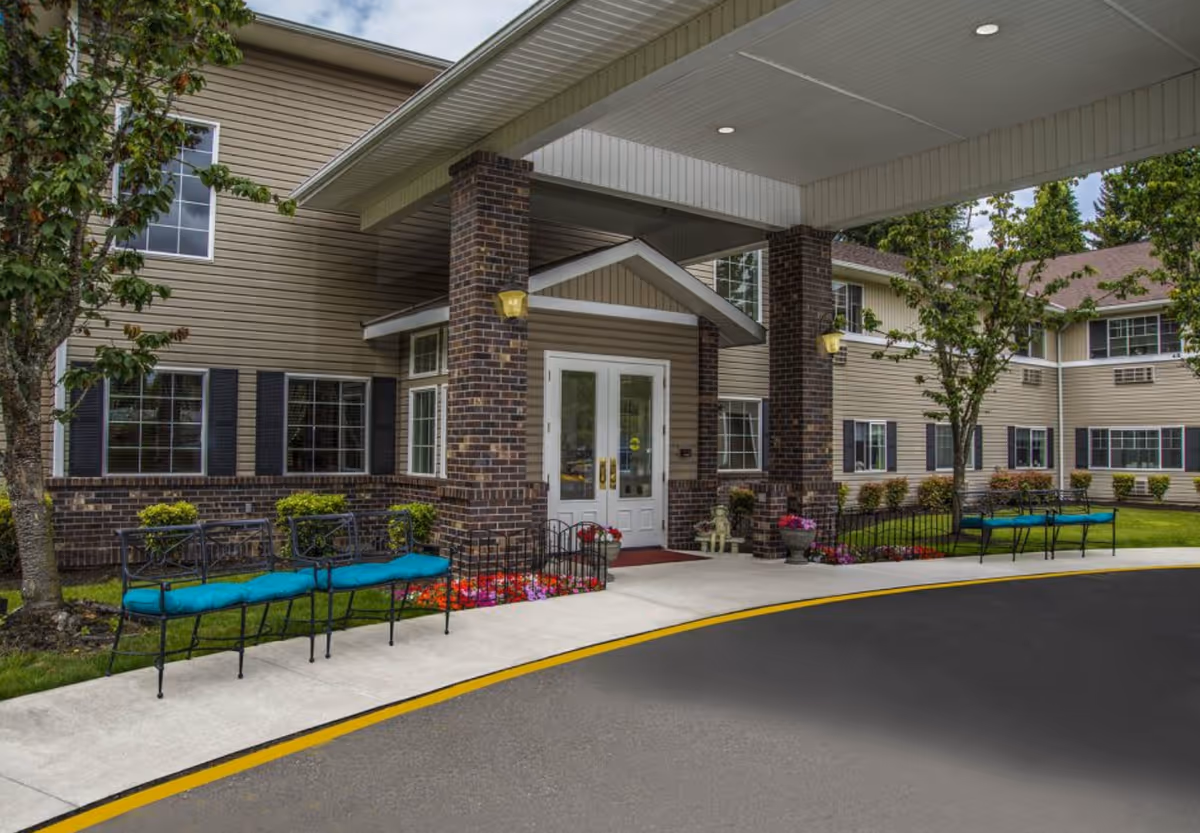 Entrance of Normandy Park Senior Living facility showing a covered drop-off area with brick pillars, double white doors, benches with blue cushions, landscaped flower beds, and trees on a sunny day.
