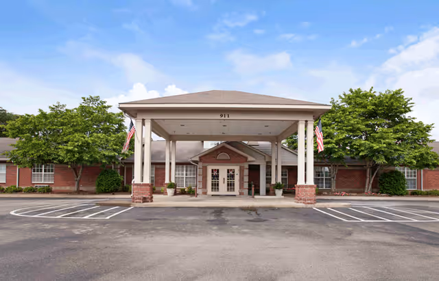 Front exterior view of a single-story brick building with a covered entrance supported by white columns. The building has several windows and is flanked by green trees. Two American flags are displayed near the entrance. The number 911 is visible above the entrance.