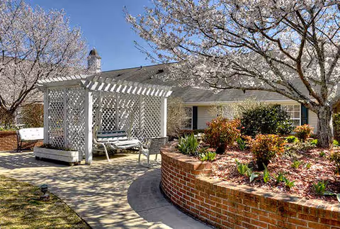Outdoor garden area with a white lattice pergola swing bench, curved brick planter filled with plants and shrubs, and blossoming trees under a clear blue sky at a senior living facility.