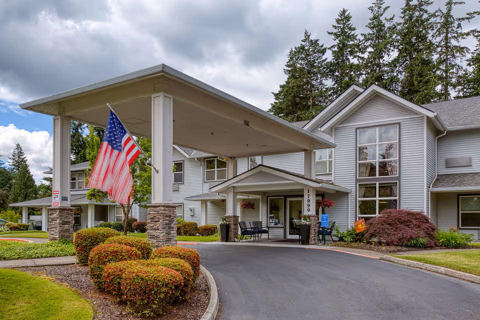 Exterior view of Homewood Assisted Living facility showing the entrance with a covered drop-off area supported by stone pillars. An American flag is displayed on one pillar. The building is light gray with large windows and surrounded by well-maintained landscaping including bushes and trees under a cloudy sky.
