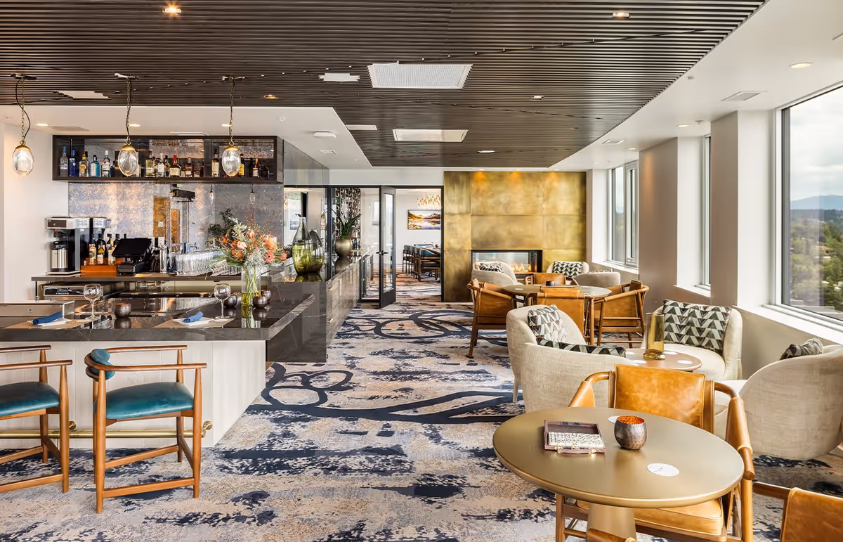 A modern and elegant lounge area with large windows offering a scenic view. The space features a bar with stools, a coffee machine, and shelves stocked with bottles. There are several seating arrangements including armchairs and small tables, a patterned carpet, and a decorative golden wall with a fireplace. The ceiling has wooden slats with recessed lighting.