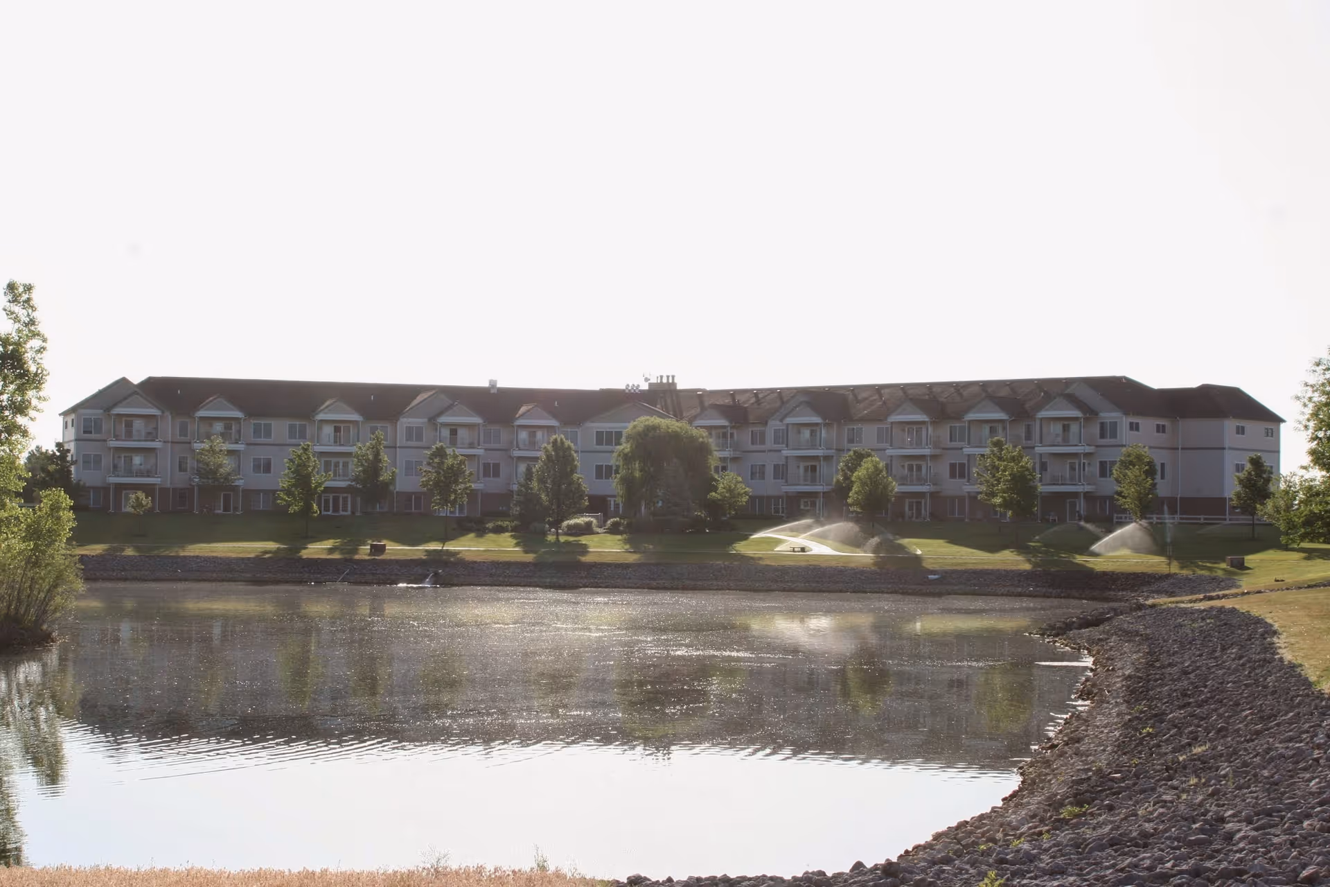 Multi-story senior living building facing a pond with fountains, trees, and a rocky shoreline.
