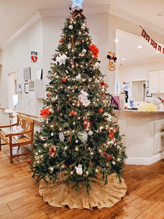 A decorated Christmas tree with white and red ornaments, lights, and a star on top, standing in a room with wooden flooring. To the left, there is a wooden bench with a cushion, and in the background, there is a counter with various items and a 'Happy Holidays' banner hanging above.