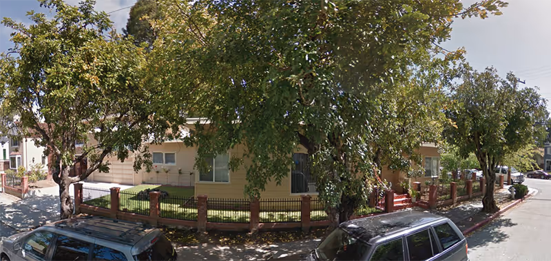 Exterior view of a single-story residential care home building surrounded by a brick and metal fence with trees providing shade. Two parked cars are visible on the street in front of the building.