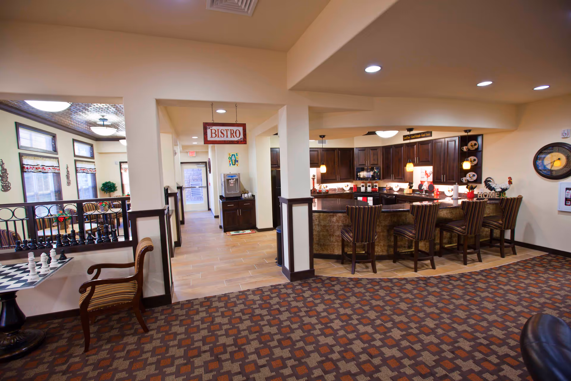 Interior view of a senior living facility showing a bistro area with a kitchen counter and bar stools. To the left, there is a seating area with a chessboard on a table and a wooden chair. The space is well-lit with ceiling lights and has a patterned carpet and tiled floor. A sign hanging from the ceiling reads 'BISTRO'.