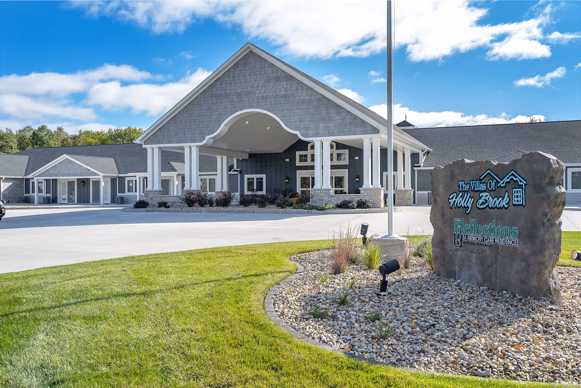 Exterior view of Villas of Holly Brook Assisted Living & Memory Care facility in Mt Carmel, IL, showing a large covered entrance with white pillars, a stone sign with the facility name, and a well-maintained lawn and landscaping under a partly cloudy blue sky.