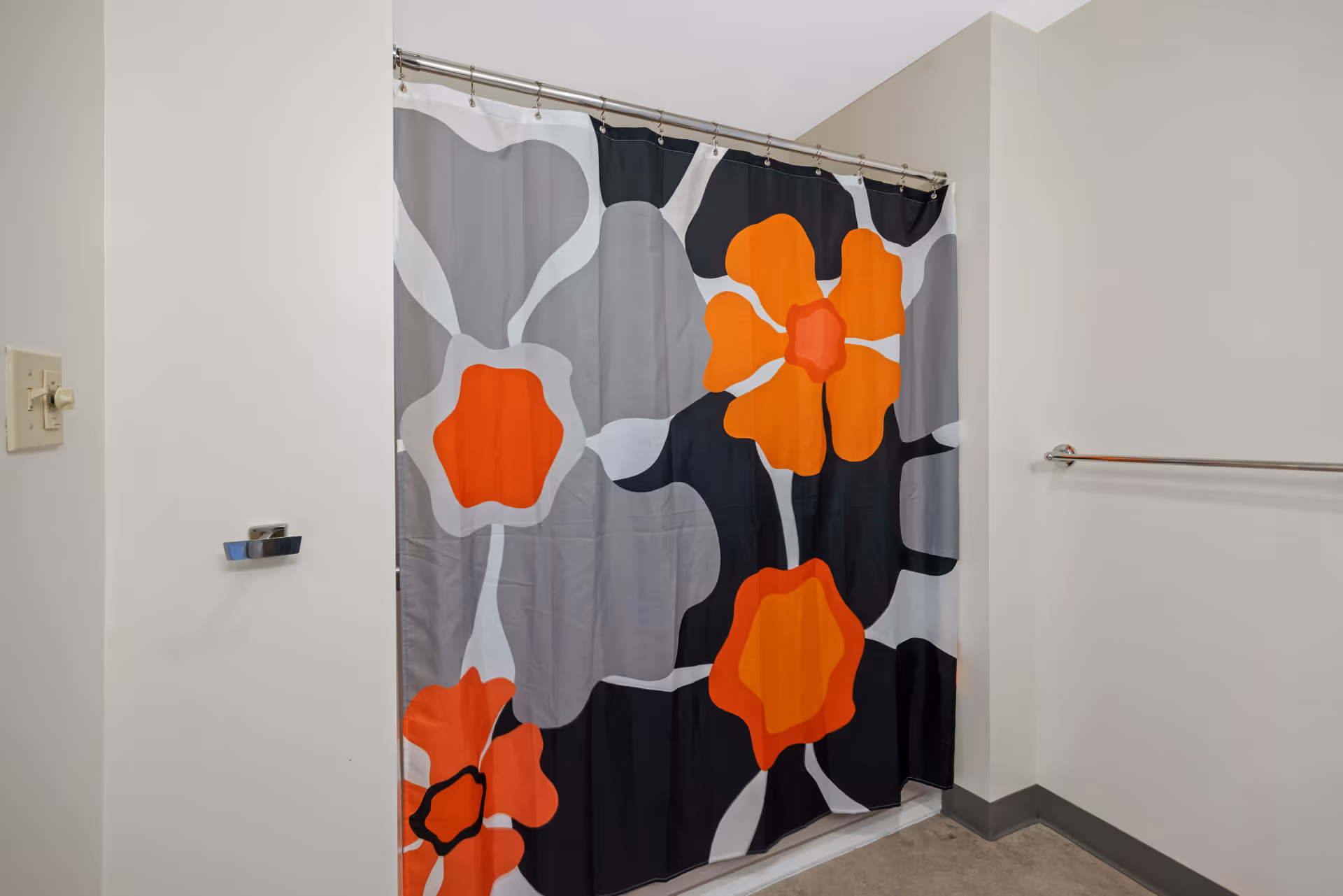 A bathroom with a colorful shower curtain featuring large abstract flowers in orange, gray, black, and white. The walls are white, and there is a towel bar on the right side and a light switch on the left wall.