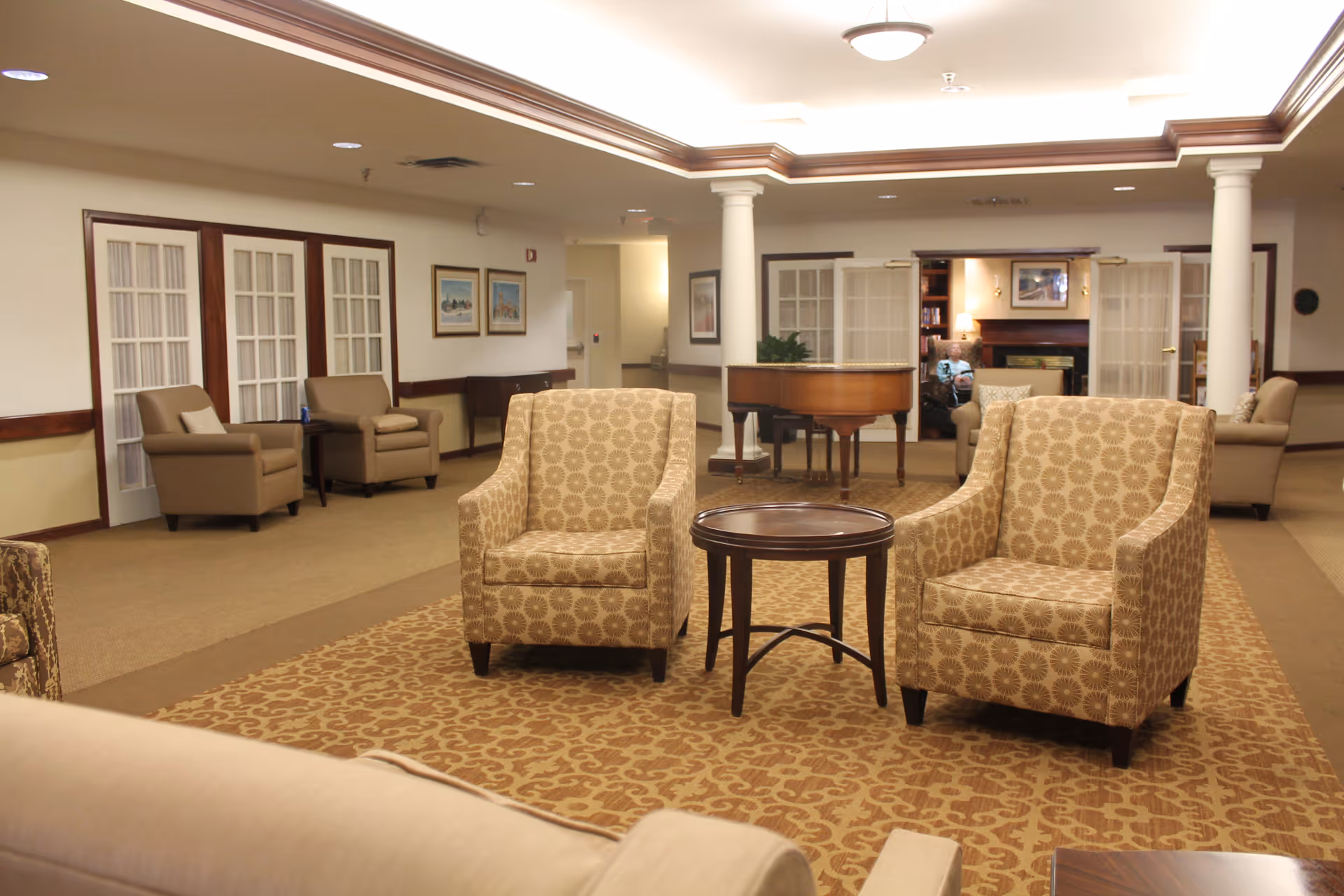 Spacious lounge with two patterned armchairs and a small round table facing a piano and additional seating in a senior living facility common area.