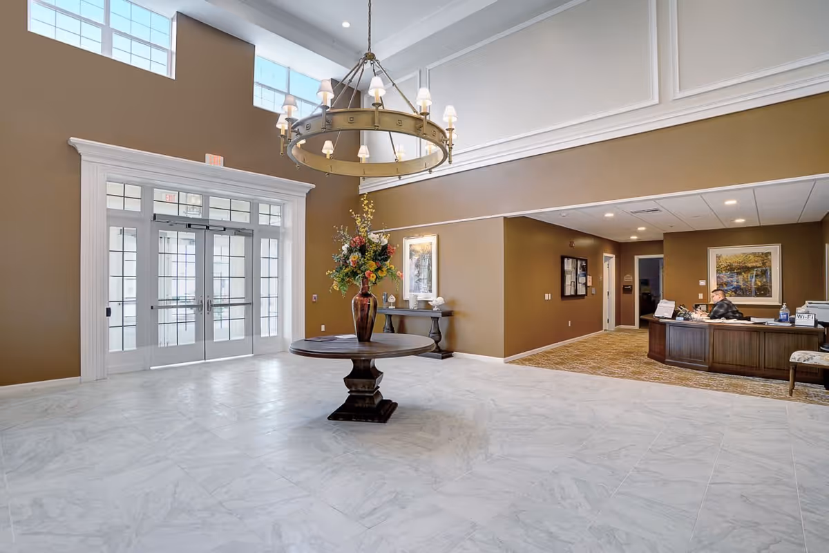 Spacious senior living facility lobby with high ceilings, large windows, a round wooden table with a tall vase of colorful flowers in the center, a chandelier hanging above, and a reception desk with a person seated behind it to the right.