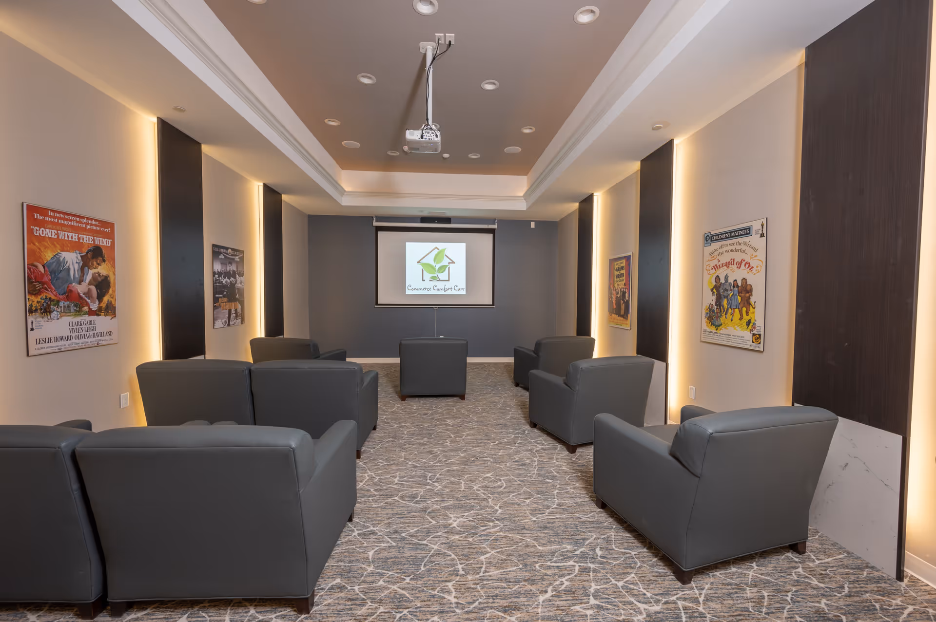 A small theater room with eight gray armchairs arranged in two rows facing a projector screen. The walls have vertical dark panels with backlighting and vintage movie posters including 'Gone with the Wind' and 'The Wizard of Oz'. The ceiling has recessed lighting and a mounted projector.