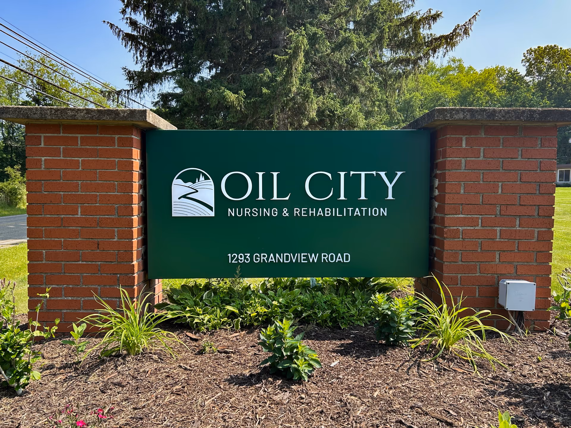 Green entrance sign mounted between two brick pillars reading 'OIL CITY NURSING & REHABILITATION 1293 GRANDVIEW ROAD' with landscaping in front and trees behind.