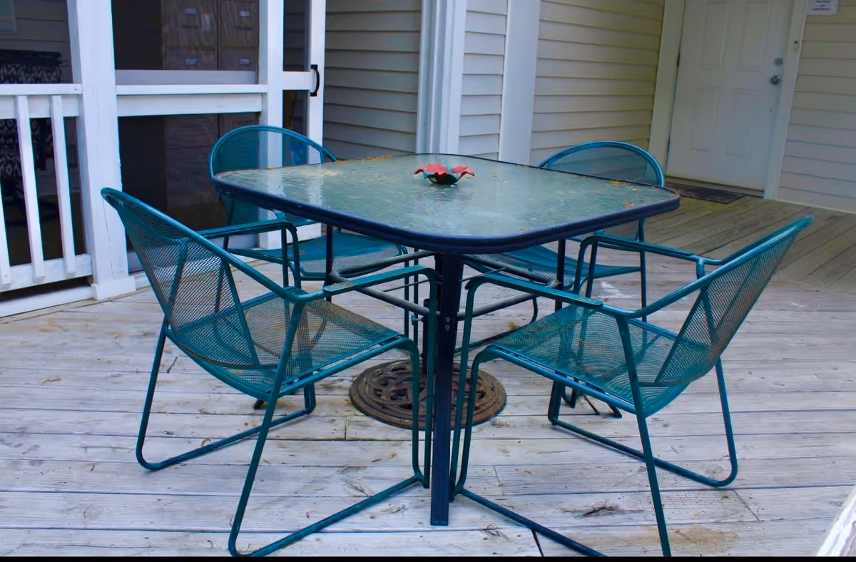 Outdoor wooden deck area with a square glass-top table and four teal metal mesh chairs arranged around it. A small decorative item is placed in the center of the table. The deck is adjacent to a building with light-colored siding and a white door.