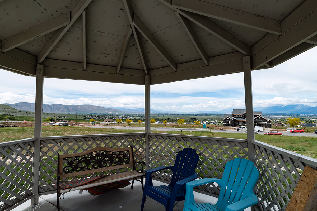 View from inside a gazebo with a wooden bench and two plastic chairs, overlooking a grassy area, a road, houses, and distant mountains under a cloudy sky.