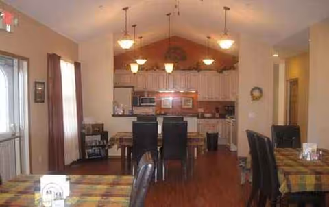 Open dining area with plaid tablecloths, dark chairs, and a kitchen in the background.