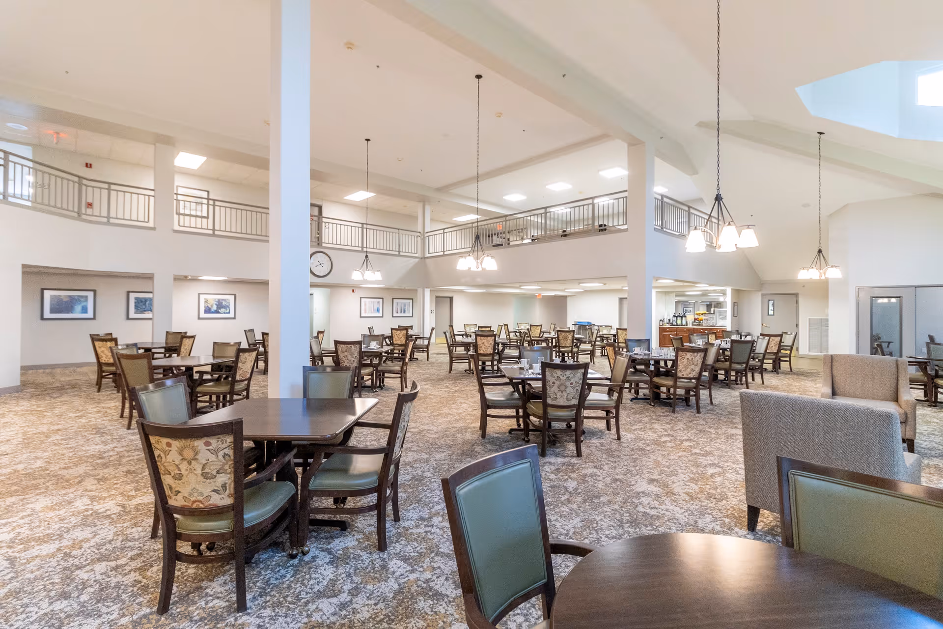 Spacious dining room with multiple tables and chairs arranged neatly. The room has high ceilings with hanging light fixtures and a second-floor balcony railing visible. The carpet has a patterned design, and there are framed pictures on the walls. A seating area with armchairs is visible on the right side.