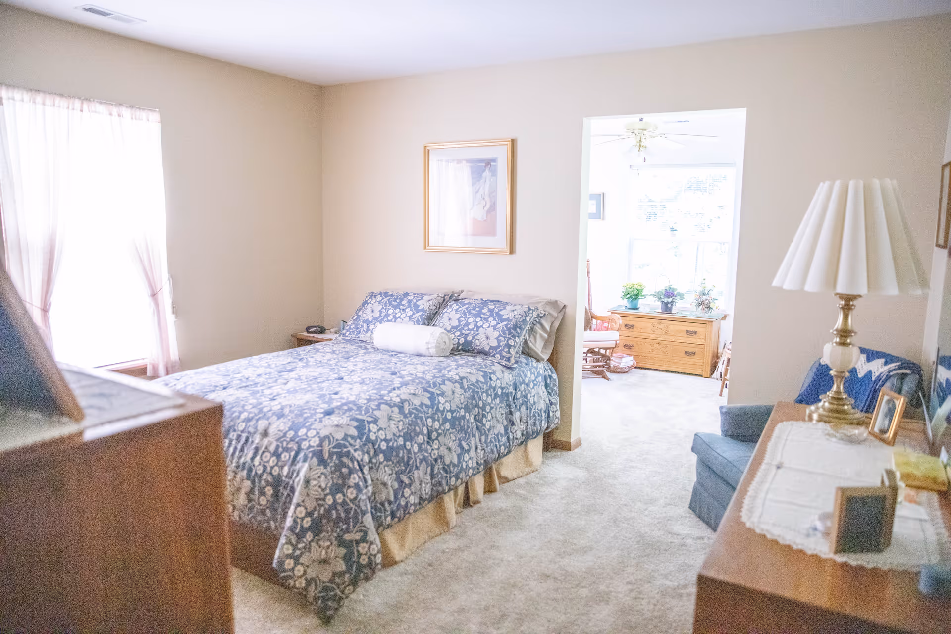 A cozy bedroom with a bed covered in a blue floral bedspread, a white bolster pillow, and a wooden nightstand with a phone. The room has beige walls, a window with sheer curtains, a framed picture on the wall, and an adjoining sitting area with a wooden dresser, plants, and a rocking chair. A wooden dresser with a white lace runner, a brass lamp, and framed photos is also visible.