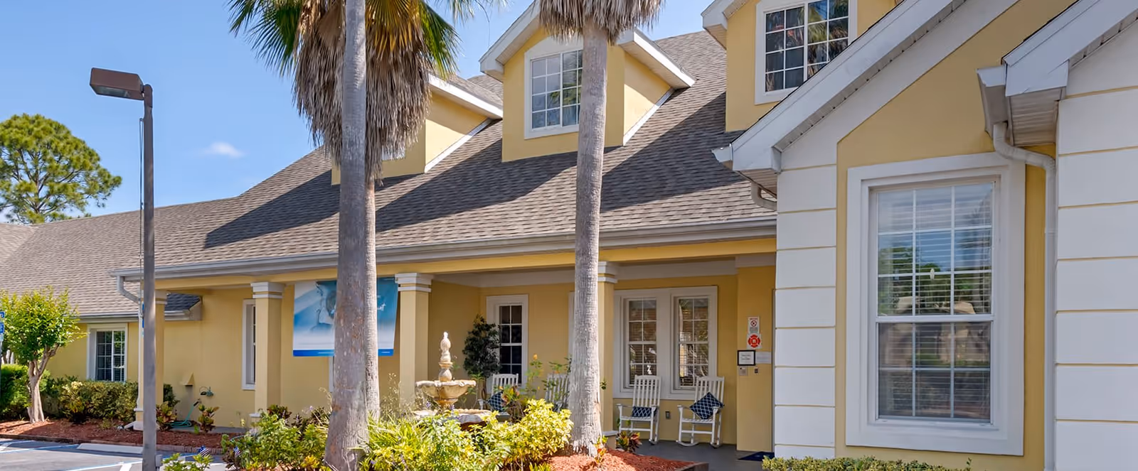 Exterior view of a yellow senior living facility building with a covered entrance, two palm trees, a small fountain, and rocking chairs on the porch under a clear blue sky.