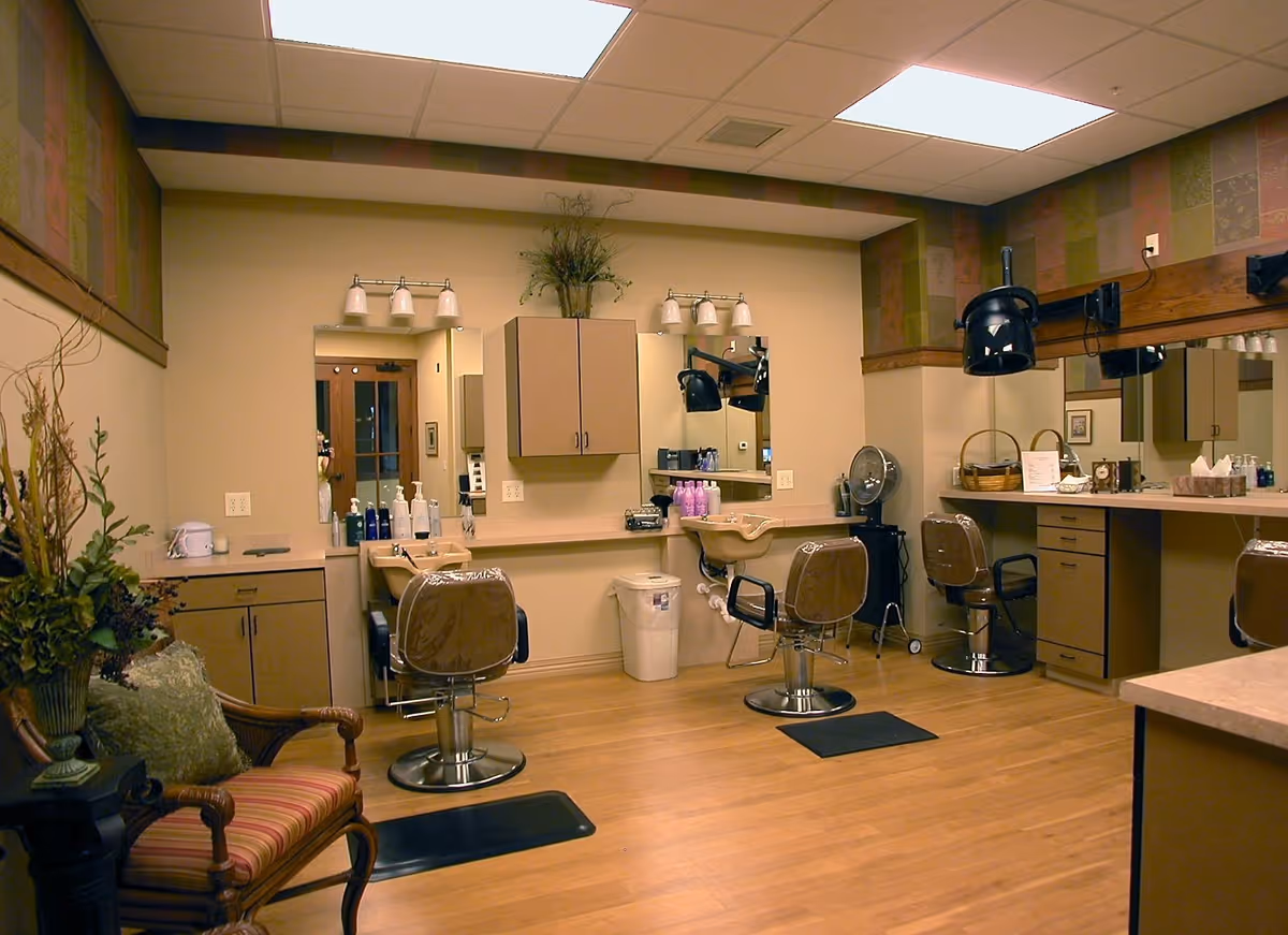Interior of a salon room with multiple styling chairs, mirrors, hair dryers, and hair care products. The room has wooden flooring, beige walls with a decorative border near the ceiling, and a comfortable armchair with a cushion and a plant in the corner.