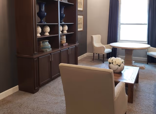 Small lounge with upholstered armchairs, a wooden coffee table topped with flowers, a round table by a window, and a dark wooden shelving unit.