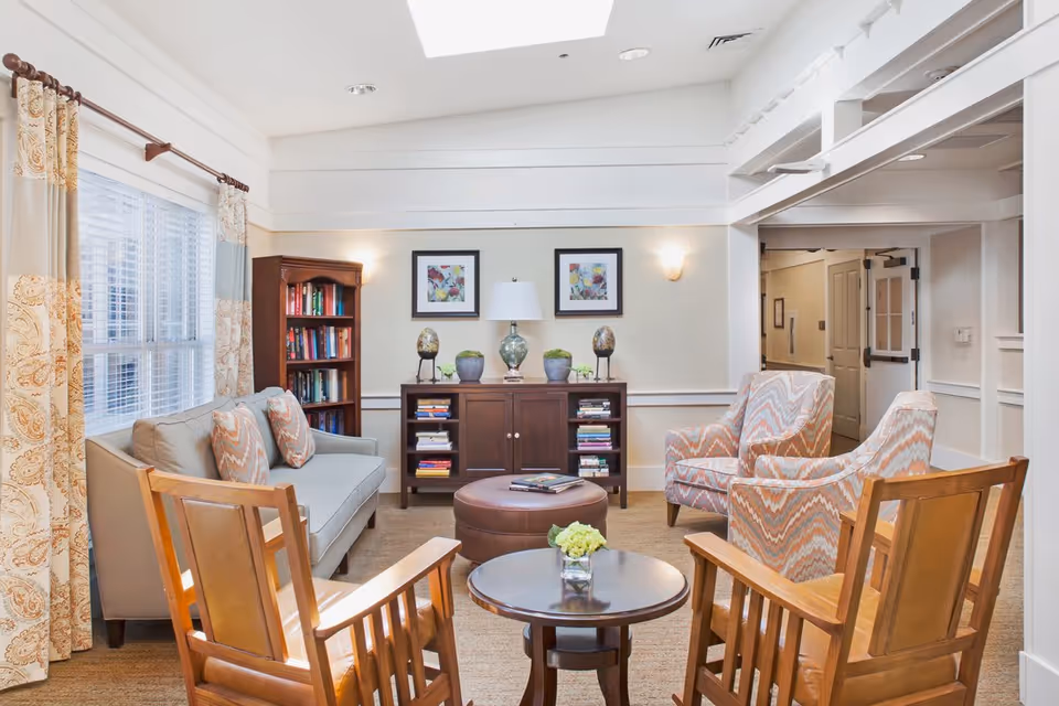 A bright communal living room with a sofa, patterned armchairs, wooden chairs, coffee tables, bookshelf and decorative artwork.
