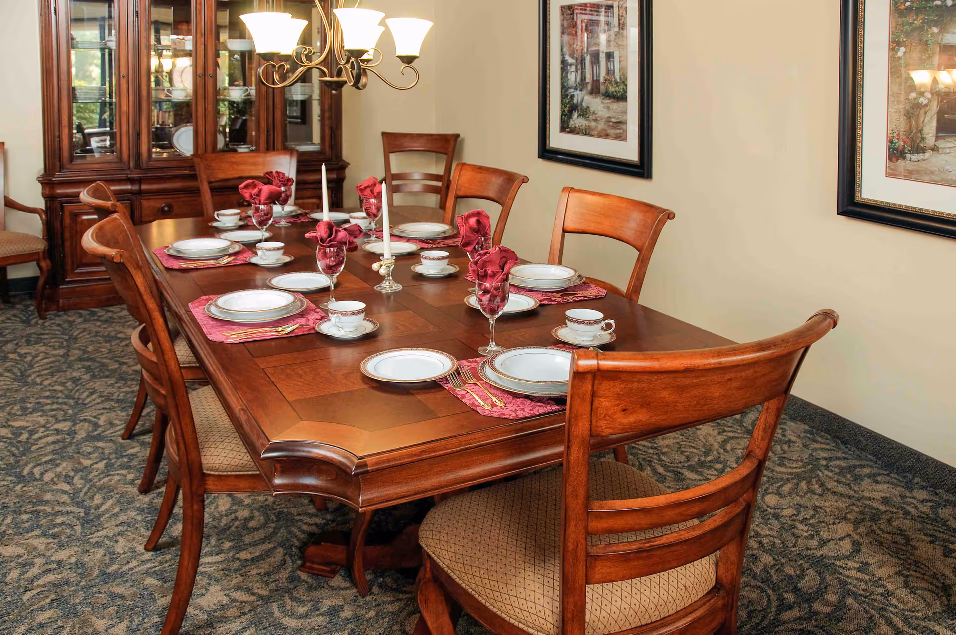 A formal dining room with a wooden dining table set for six people. The table is adorned with white plates, teacups, wine glasses with red napkins, and two white candles. The room features wooden chairs with cushioned seats, a chandelier overhead, a wooden china cabinet in the background, and framed artwork on the beige walls.