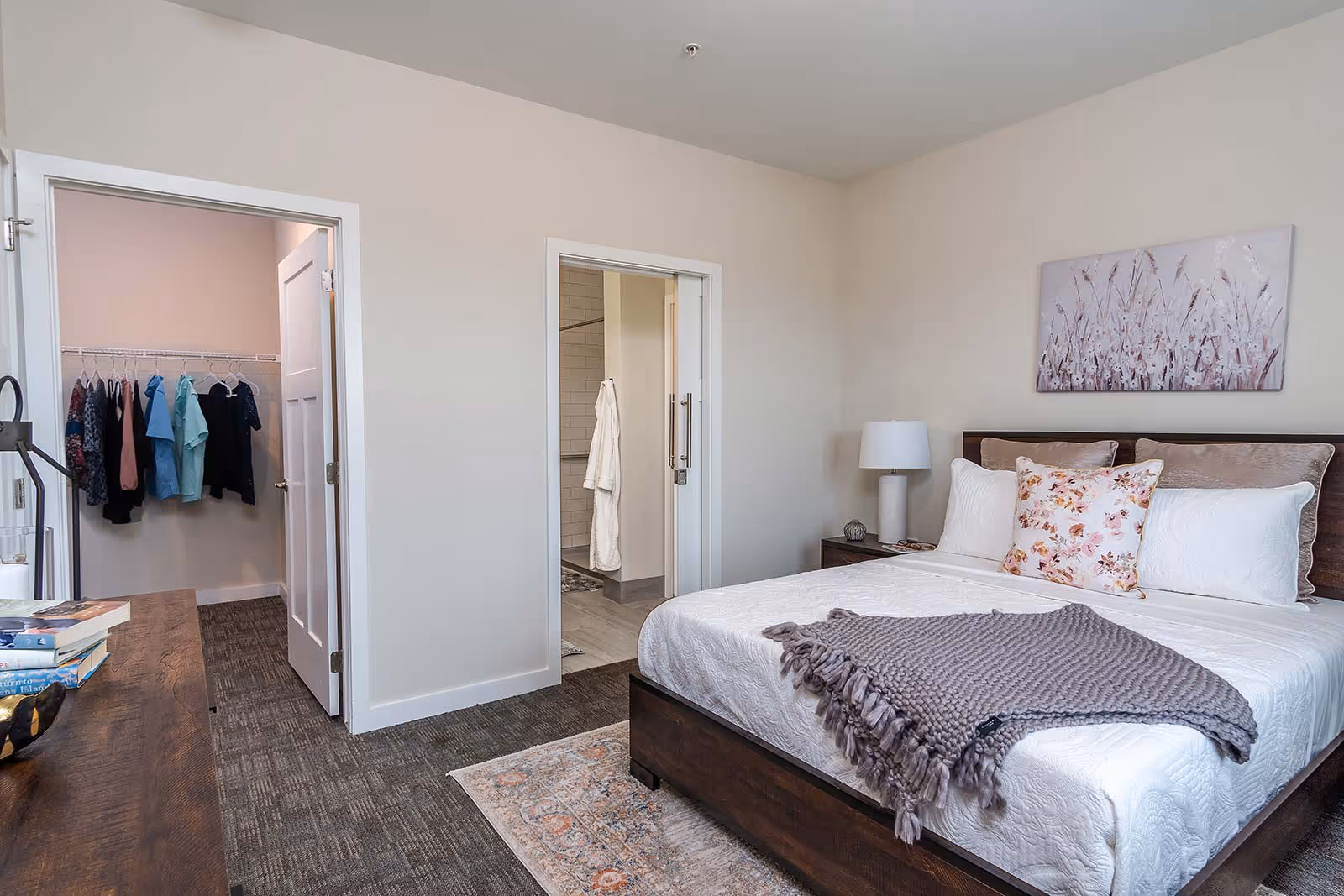 A neatly made bed with white bedding, a floral pillow, and a gray knitted throw blanket in a bedroom. To the right of the bed is a nightstand with a lamp and decorative items. On the wall above the bed is a painting of abstract flowers. To the left, there is an open door leading to a walk-in closet with clothes hanging, and another open door leading to a bathroom with a white robe hanging on the wall.