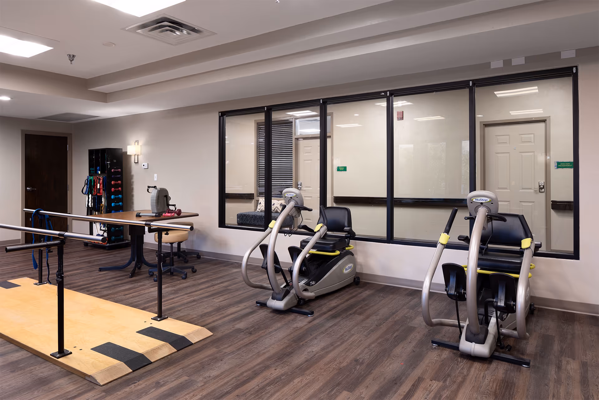 A rehabilitation or exercise room in a senior living facility with two NuStep exercise machines, parallel bars for walking support, a table with a chair, and a rack holding various exercise equipment such as dumbbells and resistance bands. The room has wood flooring and large windows looking into an adjacent room.