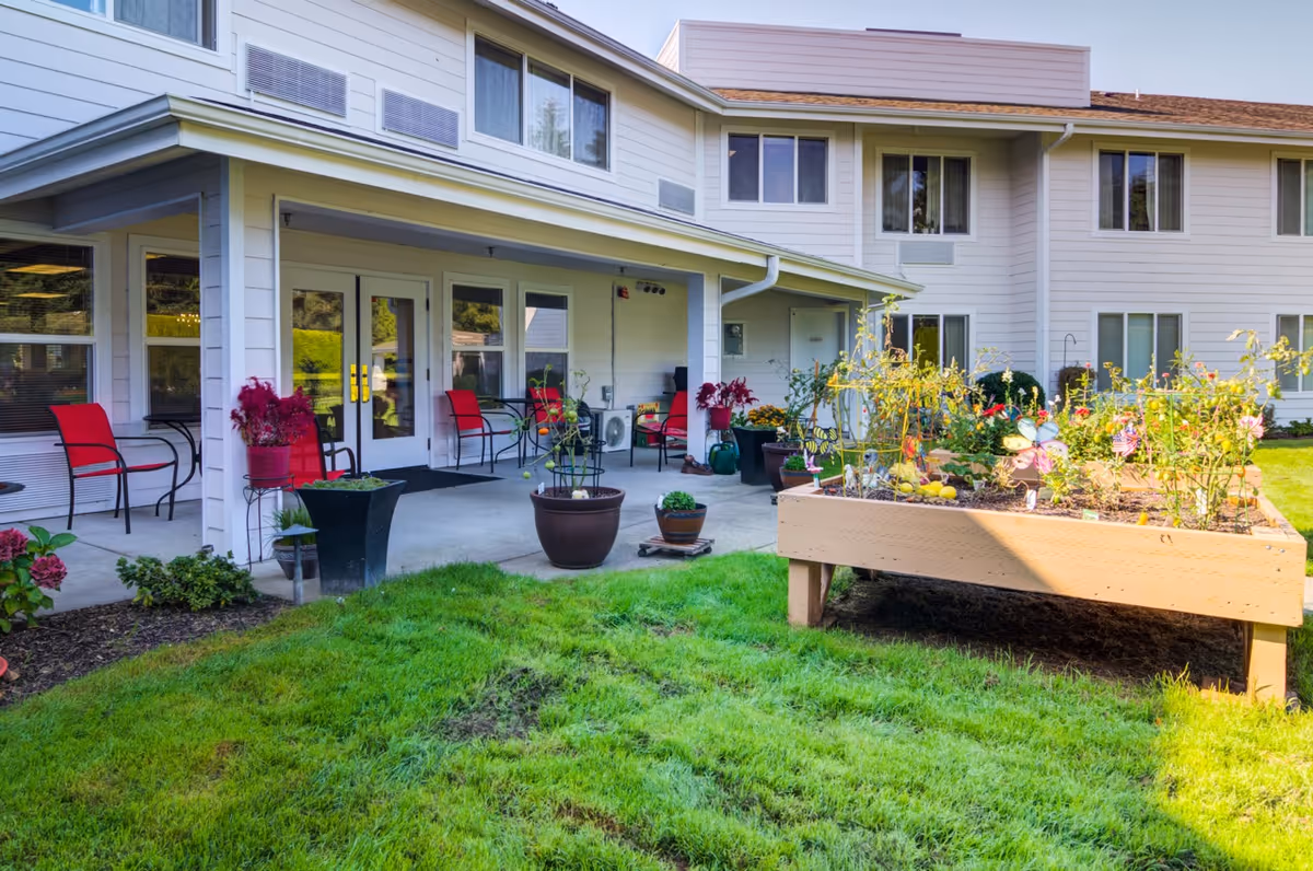 Outdoor patio area of a senior living facility with red chairs, potted plants, and a raised garden bed filled with various plants and flowers. The building has white siding and multiple windows, with a covered porch area.