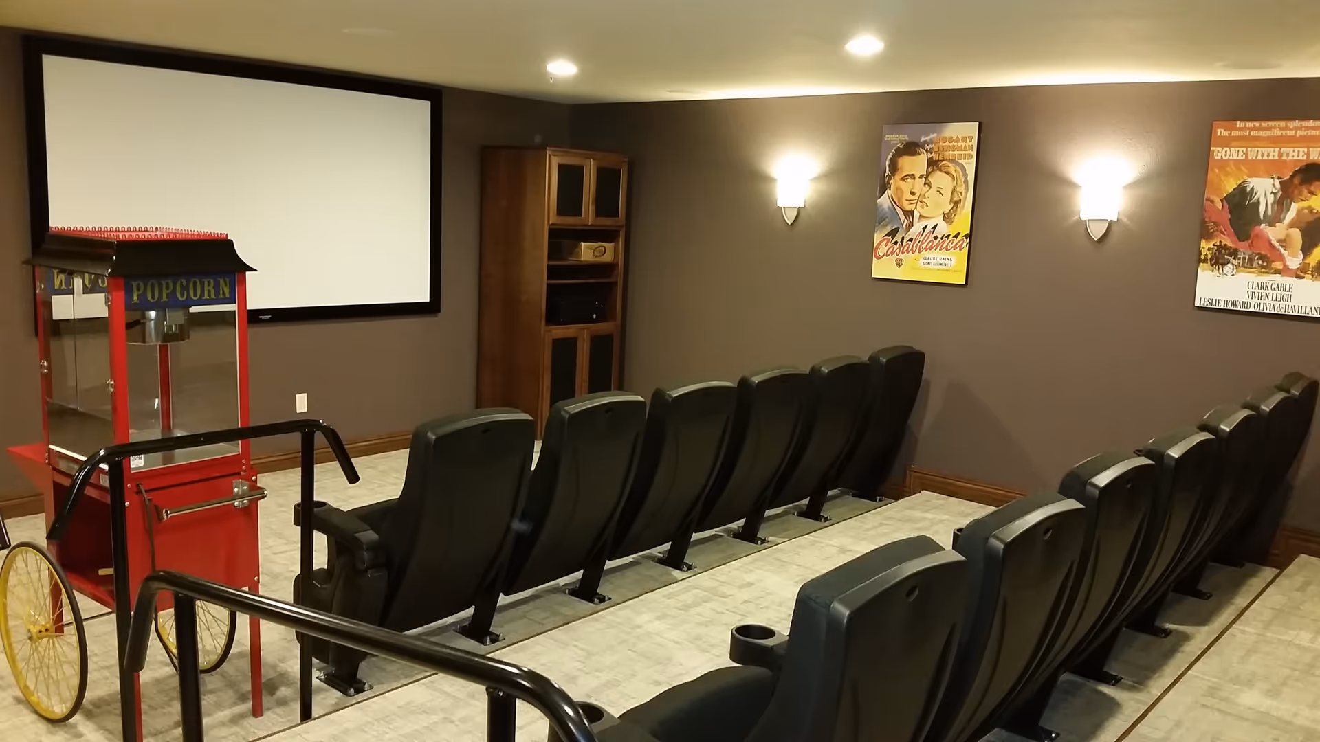 A small movie theater room with two rows of black theater seats facing a large blank projection screen. There is a red popcorn machine with yellow wheels on the left side of the room. The walls are painted brown and decorated with vintage movie posters, including Casablanca and Gone with the Wind. The room is softly lit with wall sconces.
