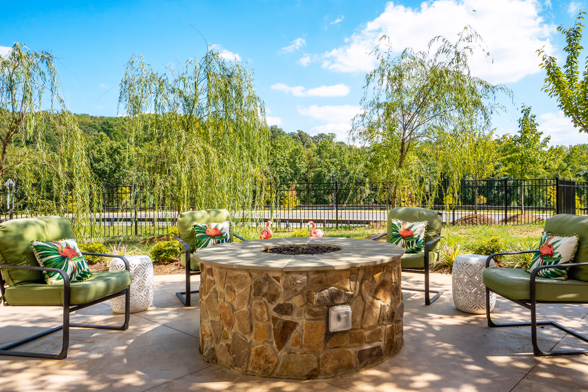 Outdoor patio area with a circular stone fire pit surrounded by four green cushioned chairs, each with a decorative pillow featuring a flamingo and tropical leaves. There are two white ceramic side tables and small pink flamingo lawn ornaments near the fire pit. The background shows a black metal fence, green trees, and a blue sky with some clouds.