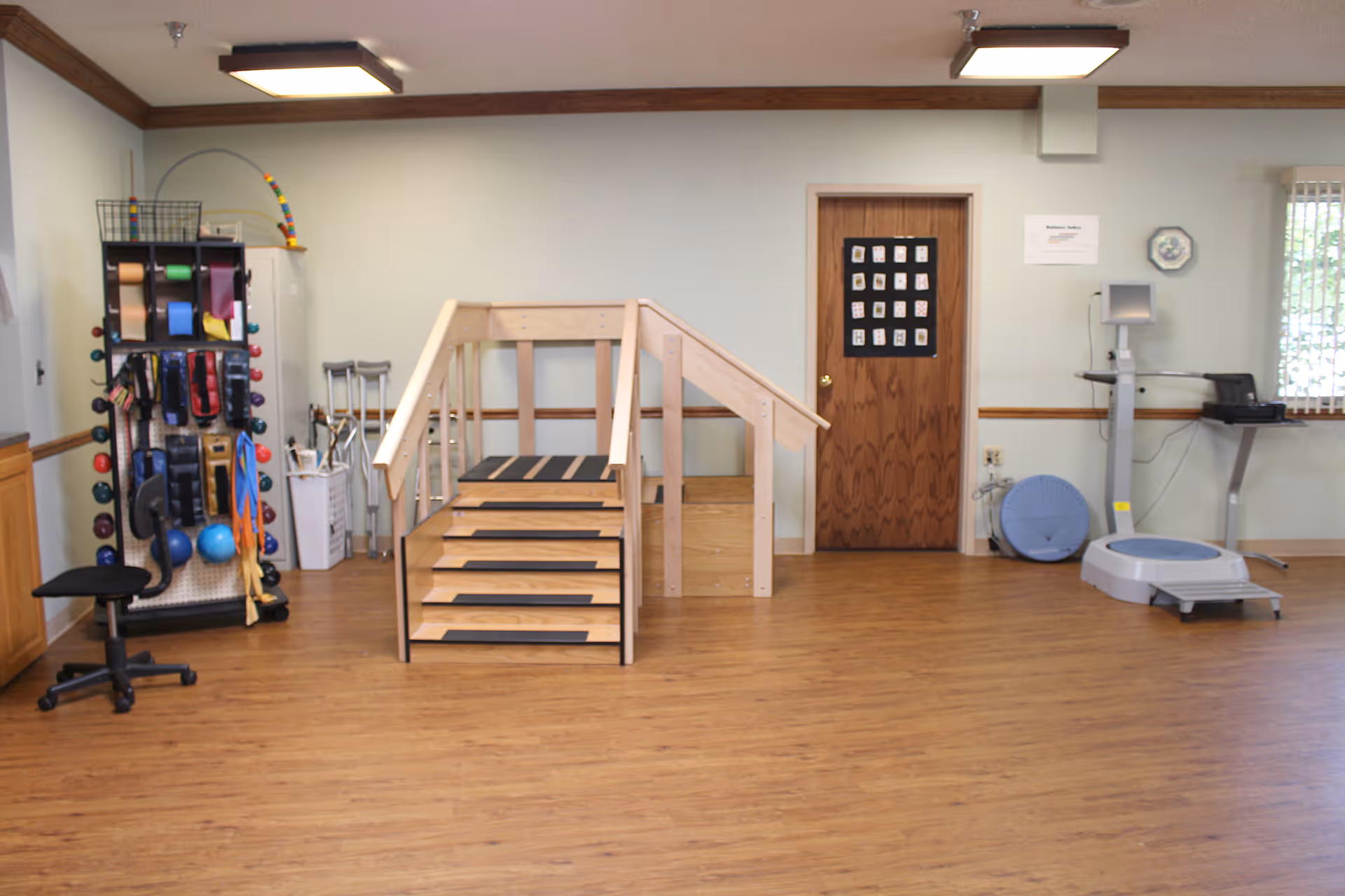 A rehabilitation or physical therapy room with wooden stairs for exercise, various therapy equipment including resistance bands, weights, exercise balls, crutches, and a balance platform. The room has wood flooring, light-colored walls, and a wooden door with a chart on it.