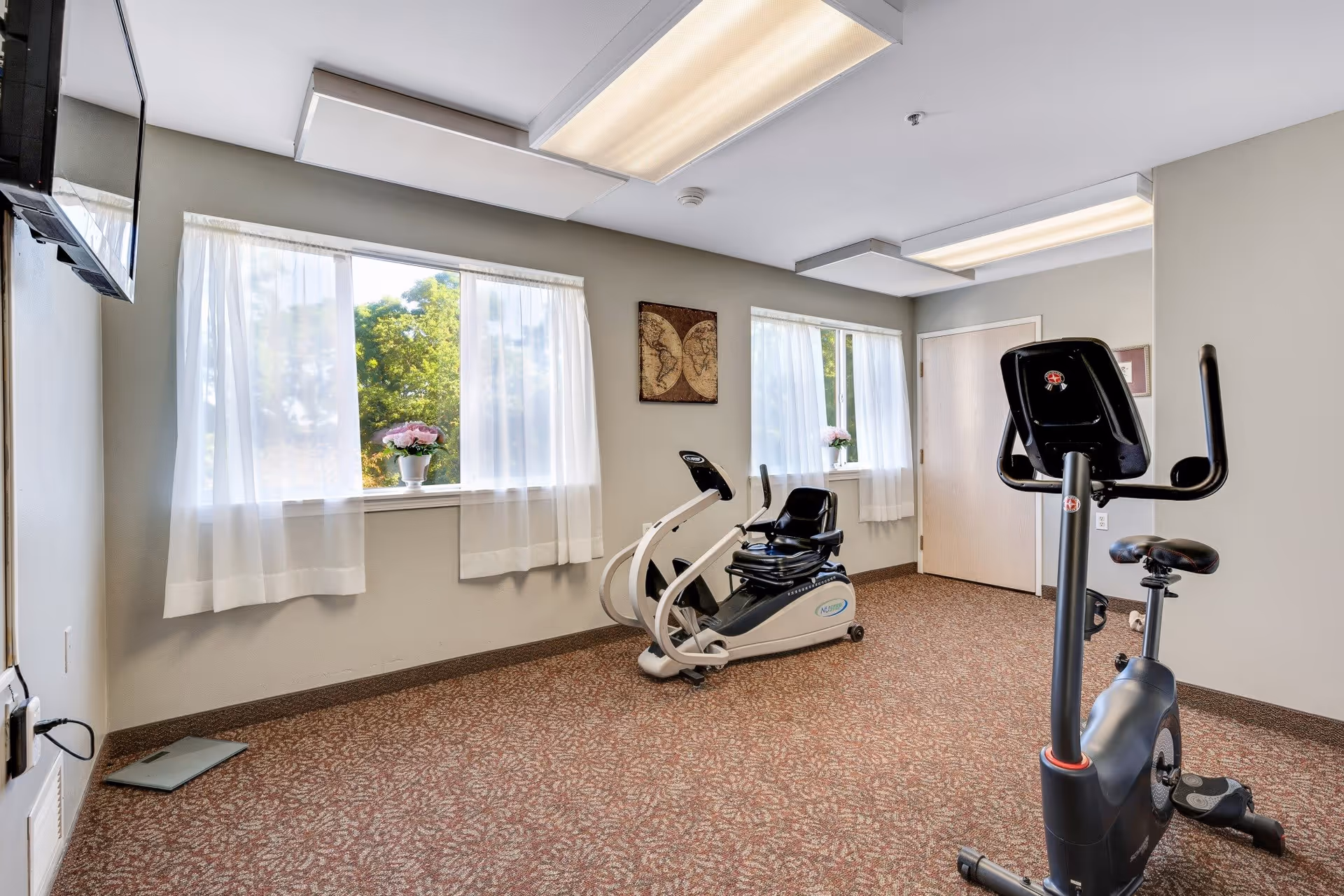 A small exercise room with two stationary bikes, beige walls, carpeted floor, two windows with sheer white curtains, a wall-mounted TV, and a decorative wall hanging between the windows.