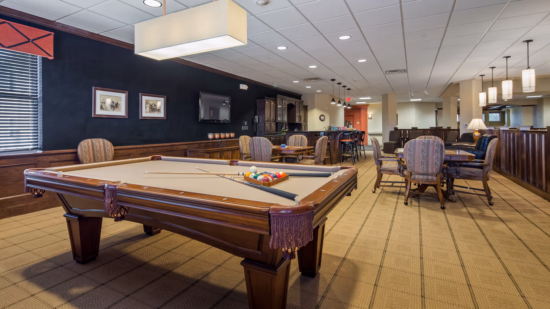 A spacious recreational room featuring a pool table with cues and balls arranged on it. The room has several upholstered chairs and tables, a wall-mounted TV, and a bar area with high stools. The lighting includes ceiling lights and hanging pendant lamps, with a window covered by blinds on the left side.