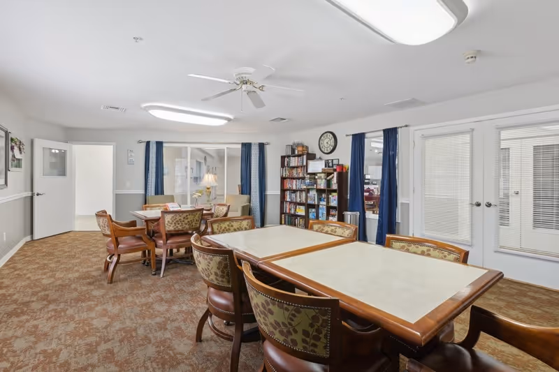A well-lit common room with two rectangular tables surrounded by cushioned chairs with floral upholstery. In the background, there is a bookshelf filled with books and games, a clock on the wall, and windows with blue curtains. The room has beige carpet flooring, white walls with a gray wainscot, and ceiling lights with a ceiling fan.