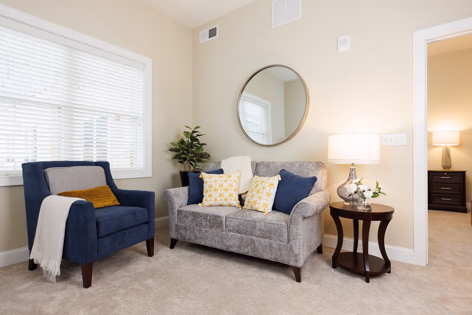 Cozy living room with a gray loveseat, blue armchair, round wall mirror, and a side table with a lamp.