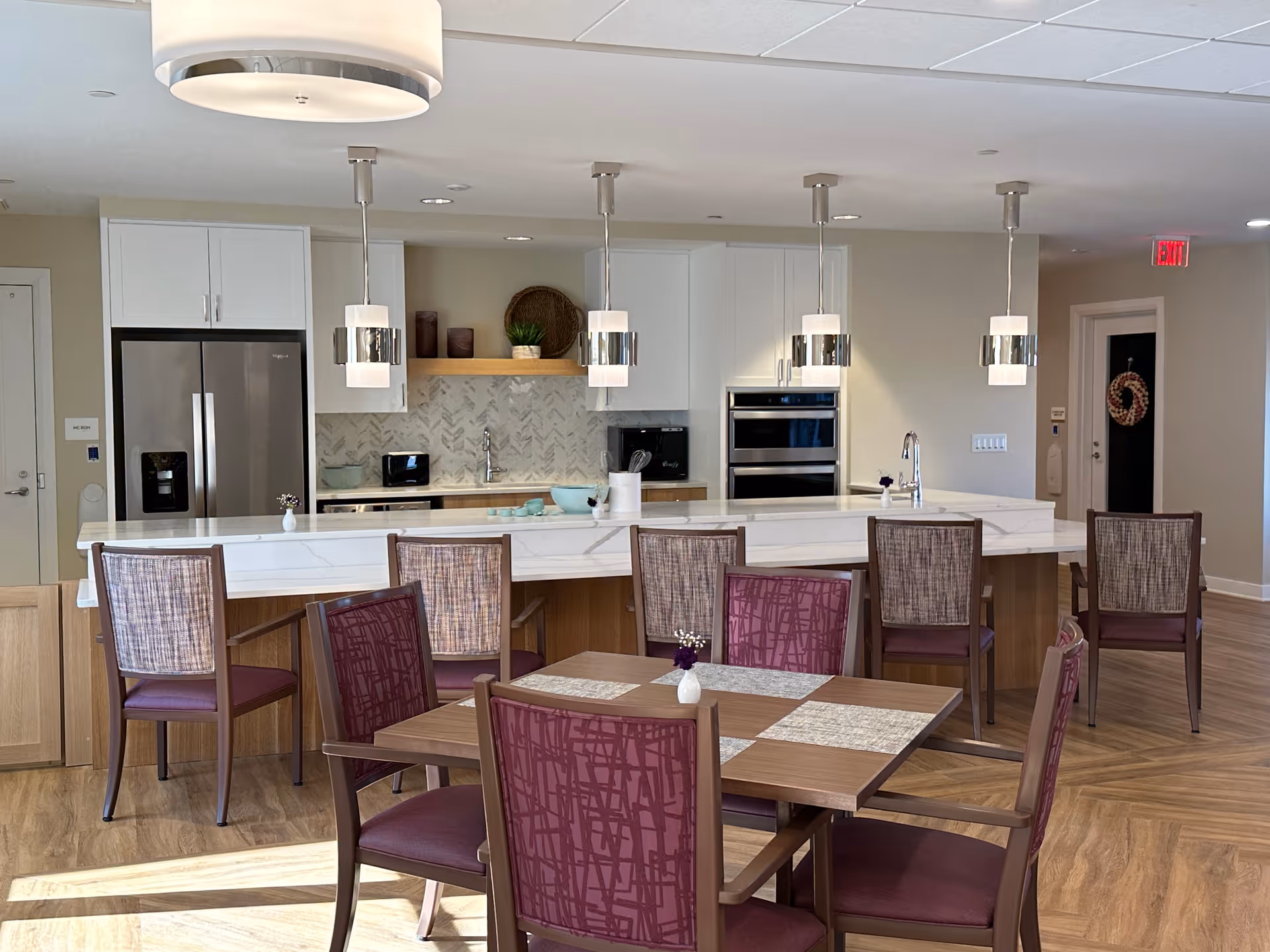 A modern kitchen and dining area in a senior living facility. The kitchen features a large white marble island with six high-back chairs, stainless steel refrigerator, built-in oven, and pendant lights hanging from the ceiling. In front of the island is a wooden dining table with four chairs upholstered in purple fabric. The floor is wood-patterned, and the walls are painted light beige. There is a door with a wreath in the background.