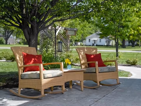Two wicker rocking chairs with beige cushions and red pillows are placed on a concrete patio under a large tree. A small wicker table with a vase of yellow flowers sits between the chairs. In the background, there is a green lawn, trees, and a white house across the street.
