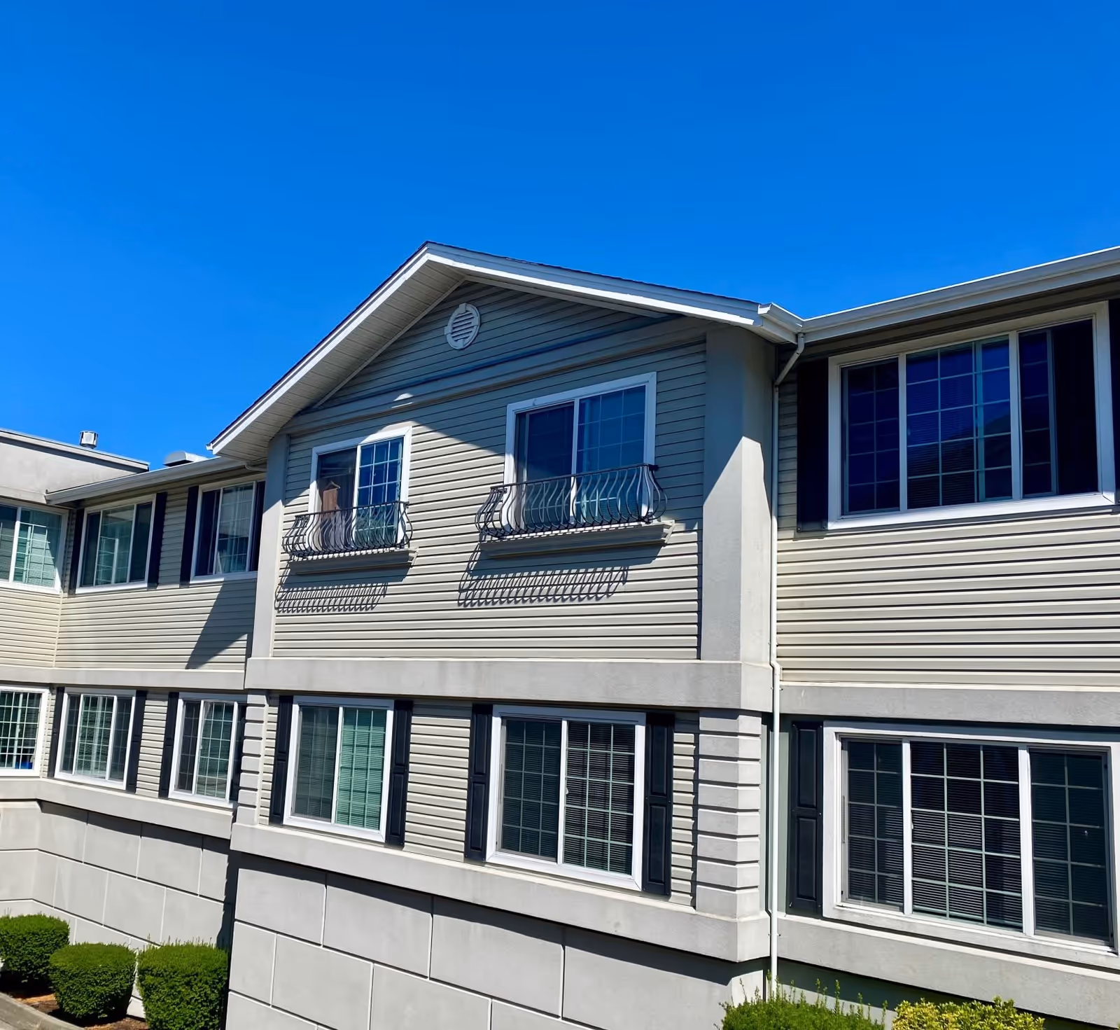 Exterior view of a two-story building with beige siding and multiple windows, some with decorative black railings, under a clear blue sky. Small green bushes are visible at the base of the building.