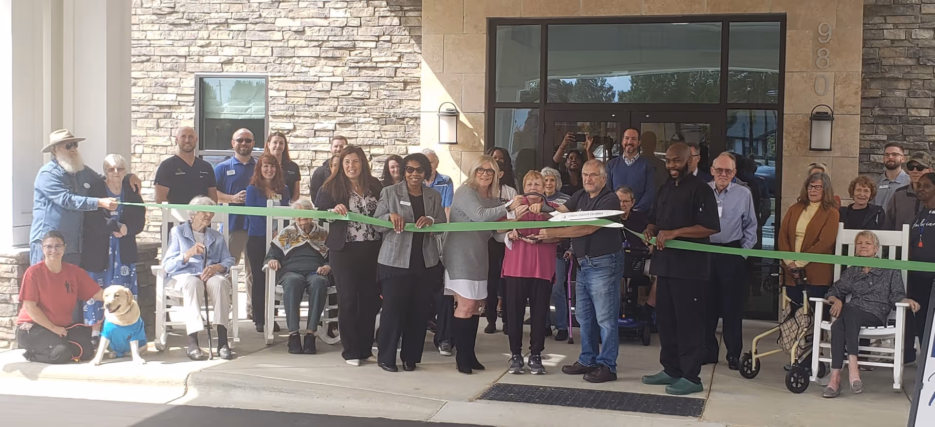 A group of people gathered outside the entrance of a building for a ribbon-cutting ceremony. Several individuals are holding a long green ribbon, and one person is cutting it with large scissors. The building has a stone facade and glass doors. Some elderly individuals are seated in rocking chairs and wheelchairs, and a dog wearing a blue vest is sitting on the ground. The atmosphere appears celebratory and community-focused.