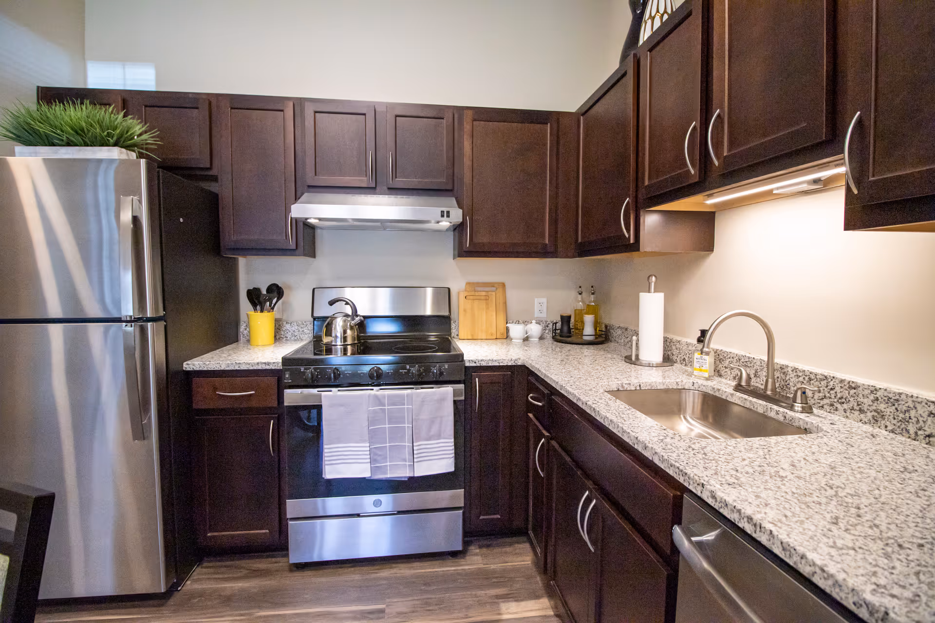 A modern kitchen with dark wood cabinets, a stainless steel refrigerator, stove with a kettle on it, granite countertops, a sink with a faucet, and various kitchen utensils and accessories including a paper towel holder and a small plant on top of the refrigerator.