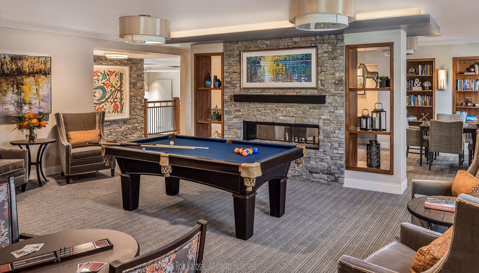 A cozy common area in a retirement community featuring a pool table with billiard balls and cues, surrounded by comfortable armchairs and small tables. The room has a stone fireplace with a colorful framed painting above it, built-in wooden shelves with decorative items, and a carpeted floor. In the background, there is a glimpse of a study or reading area with bookshelves and a table with chairs.