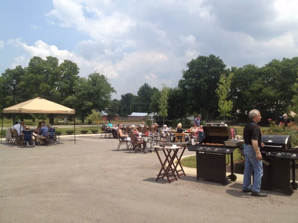 Outdoor gathering at Carrick Glen Senior Living with several seniors seated at tables under a canopy and in open air, while a man grills food on two barbecue grills. The setting is a paved area with trees and greenery in the background under a partly cloudy sky.