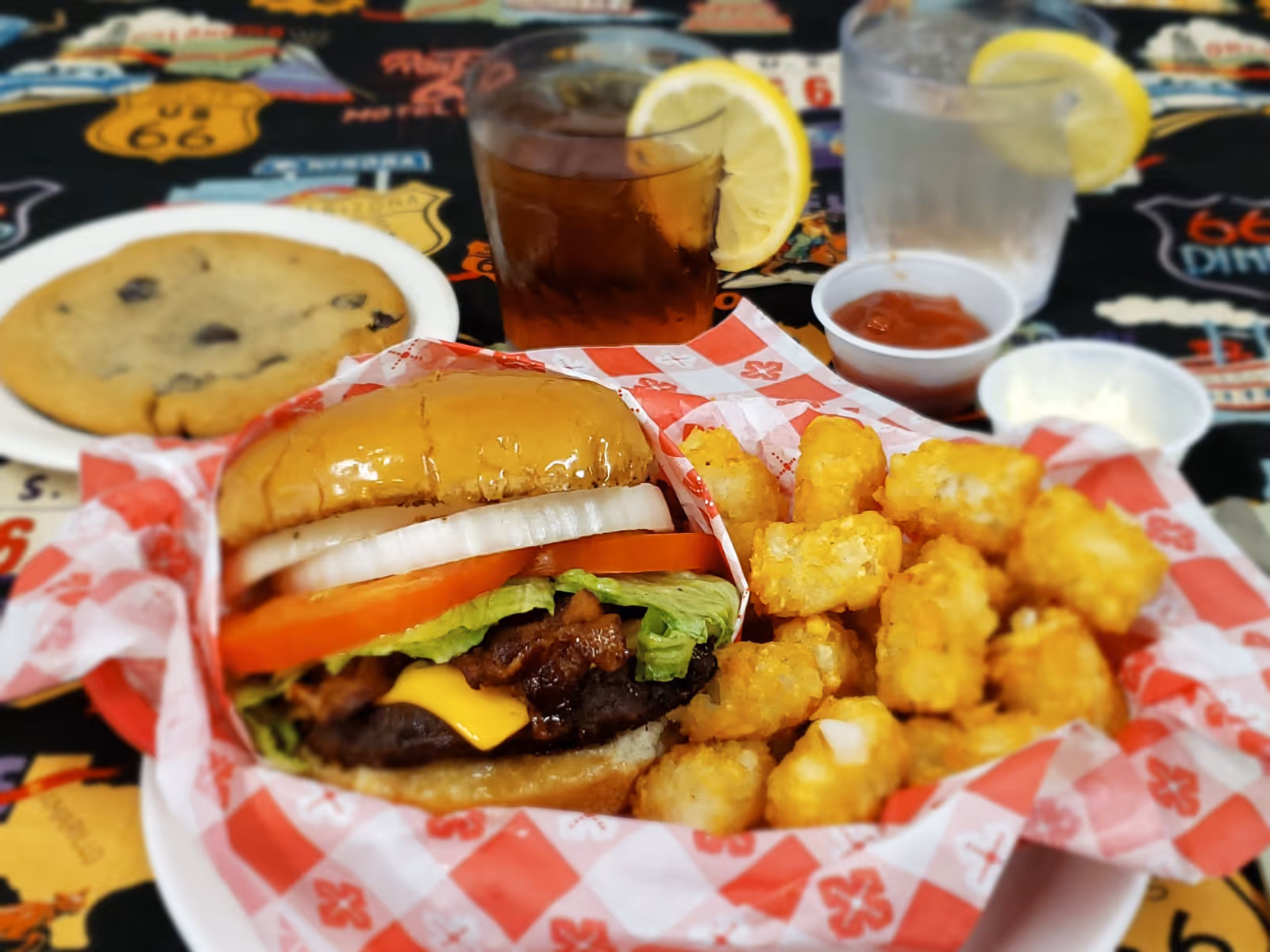 A meal consisting of a cheeseburger with lettuce, tomato, and onion, a side of tater tots, a chocolate chip cookie on a white plate, a glass of iced tea with a lemon slice, a glass of water with a lemon slice, and small containers of ketchup and mayonnaise on a table with a colorful Route 66 themed tablecloth.