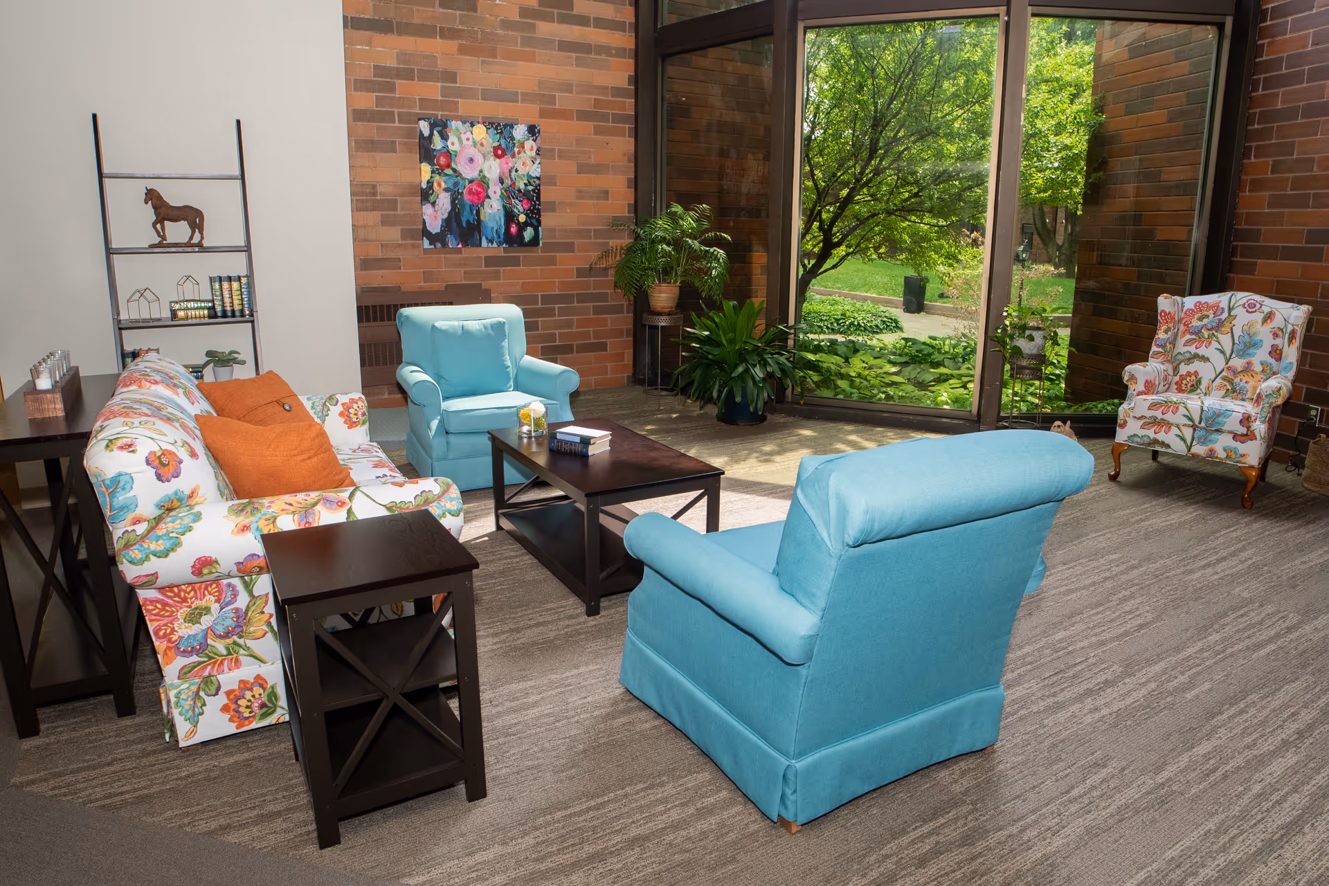 A cozy living room area with floral patterned sofas and blue armchairs arranged around a dark wooden coffee table. There is a large window showing a green garden outside, a brick wall with a colorful floral painting, and several potted plants adding a touch of nature indoors.