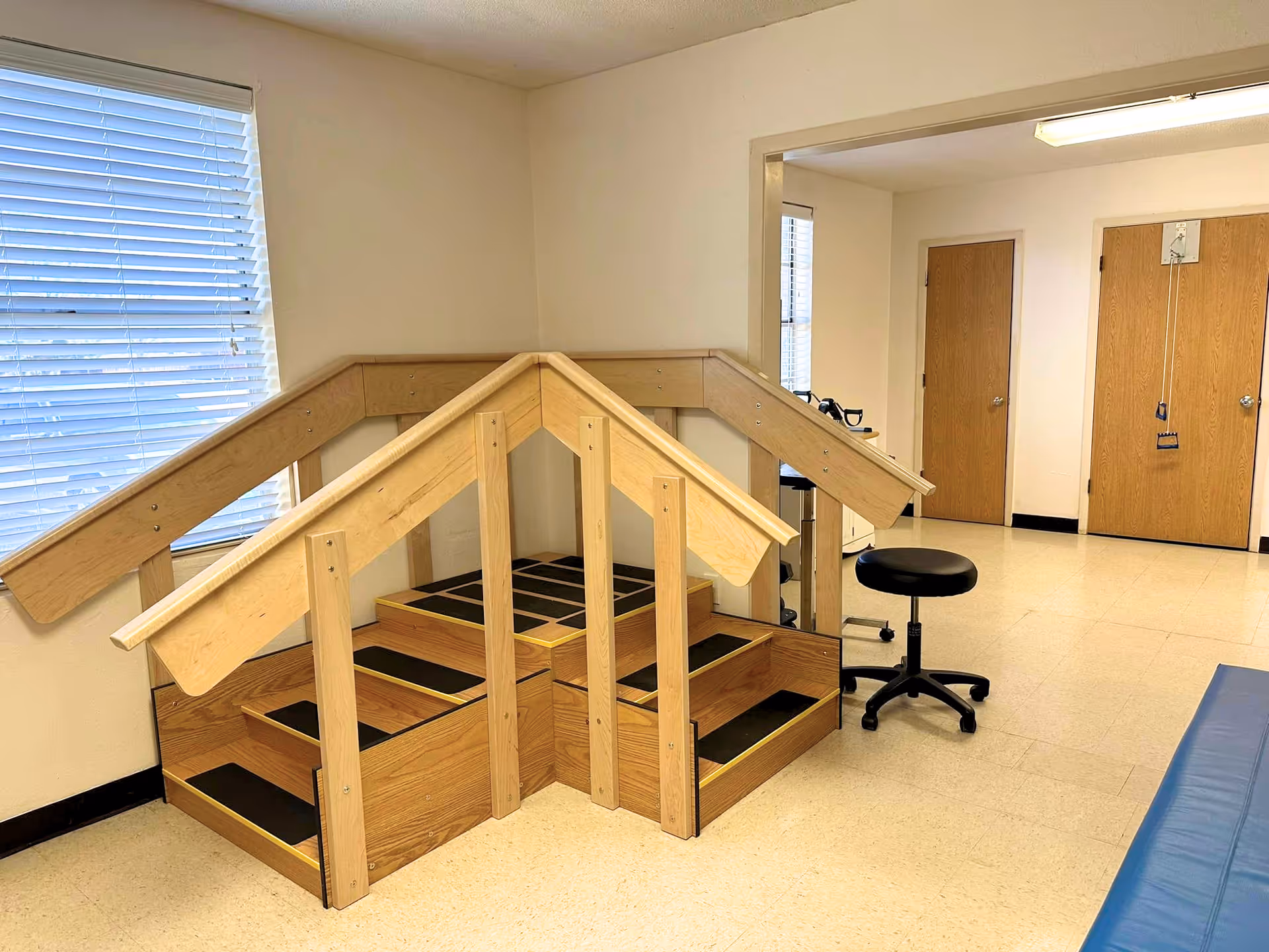 Therapy room interior with wooden practice stairs and handrails, a rolling stool, doors and a window.