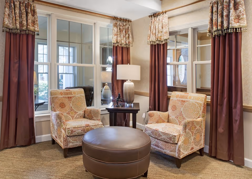 Cozy sitting area with two patterned armchairs, a round leather ottoman, and a side table with a lamp in front of curtained windows.