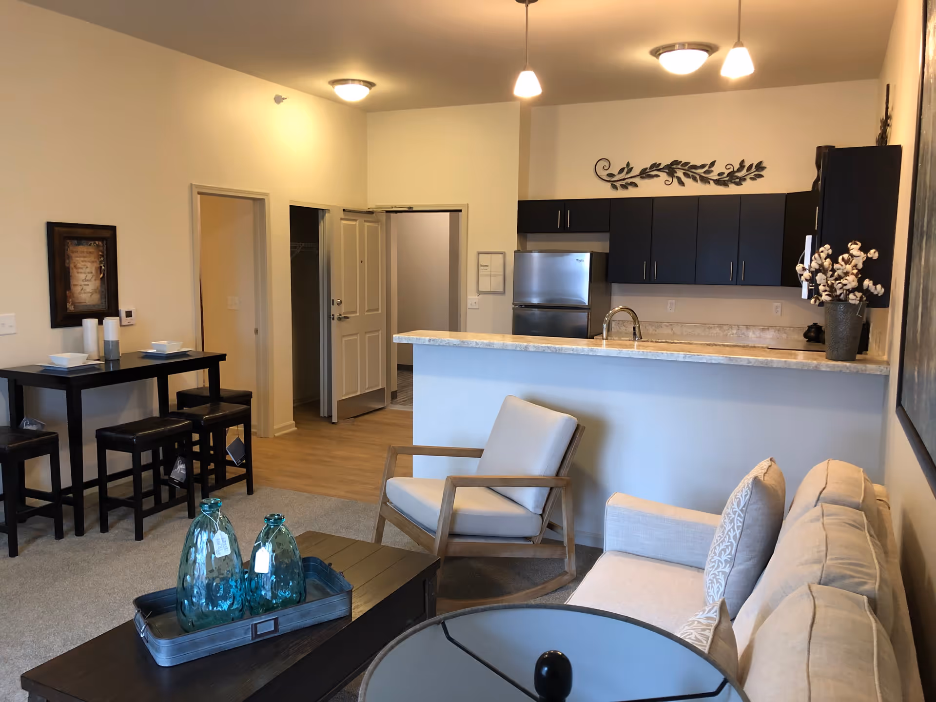 Interior view of a senior living apartment showing a living room with a beige sofa and armchair, a coffee table with decorative blue glass bottles, a small dining table with four chairs, and a kitchen area with dark cabinets, a refrigerator, and a countertop with a sink. The walls are light-colored and there is a decorative metal wall art above the kitchen cabinets.